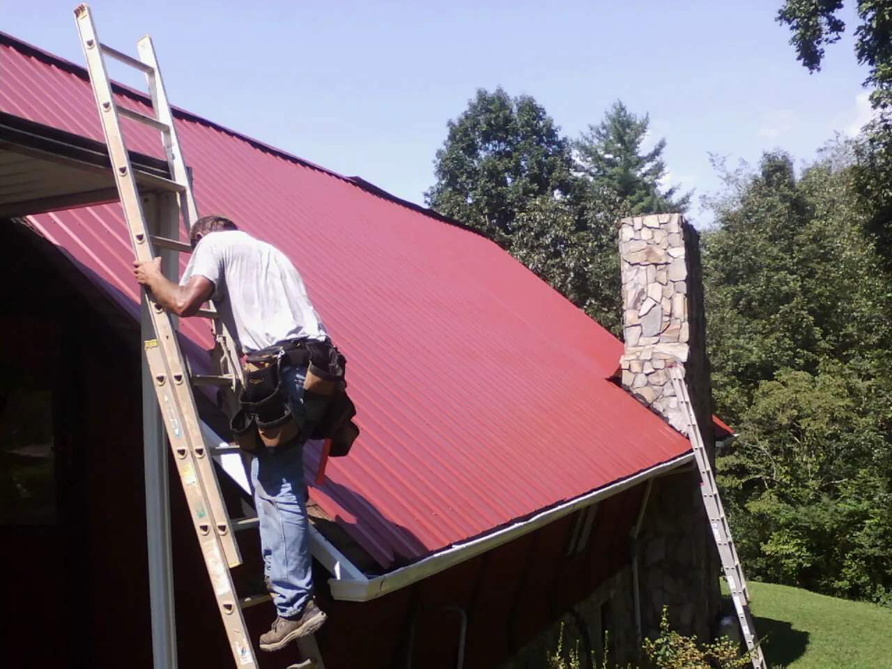 Roofer on a ladder beside a red metal roof and stone chimney — Soffit & Fascia Repair by Roofing Brothers in Hockingport